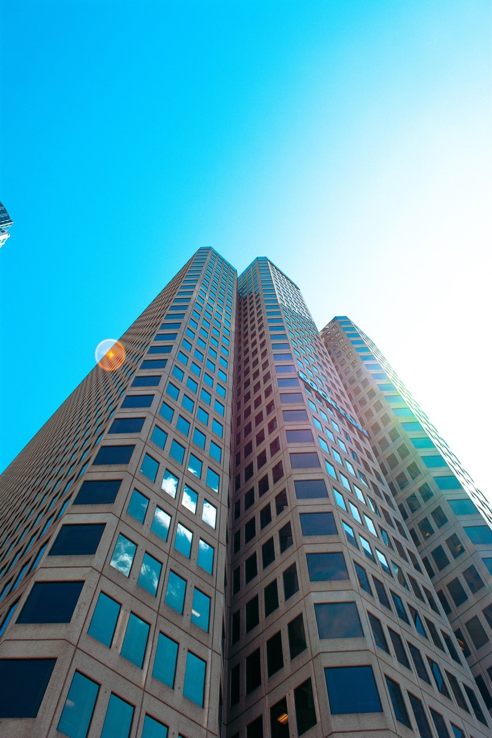 Looking straight up a faceted tan tower into blue sky, a lens flare at its edge — from the Aerial study