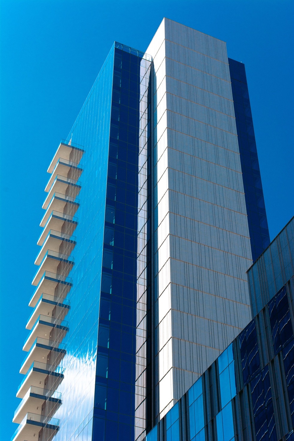 A glass tower with stacked cantilevered balconies beside a pale stone shaft, under deep blue sky — from the Aerial study