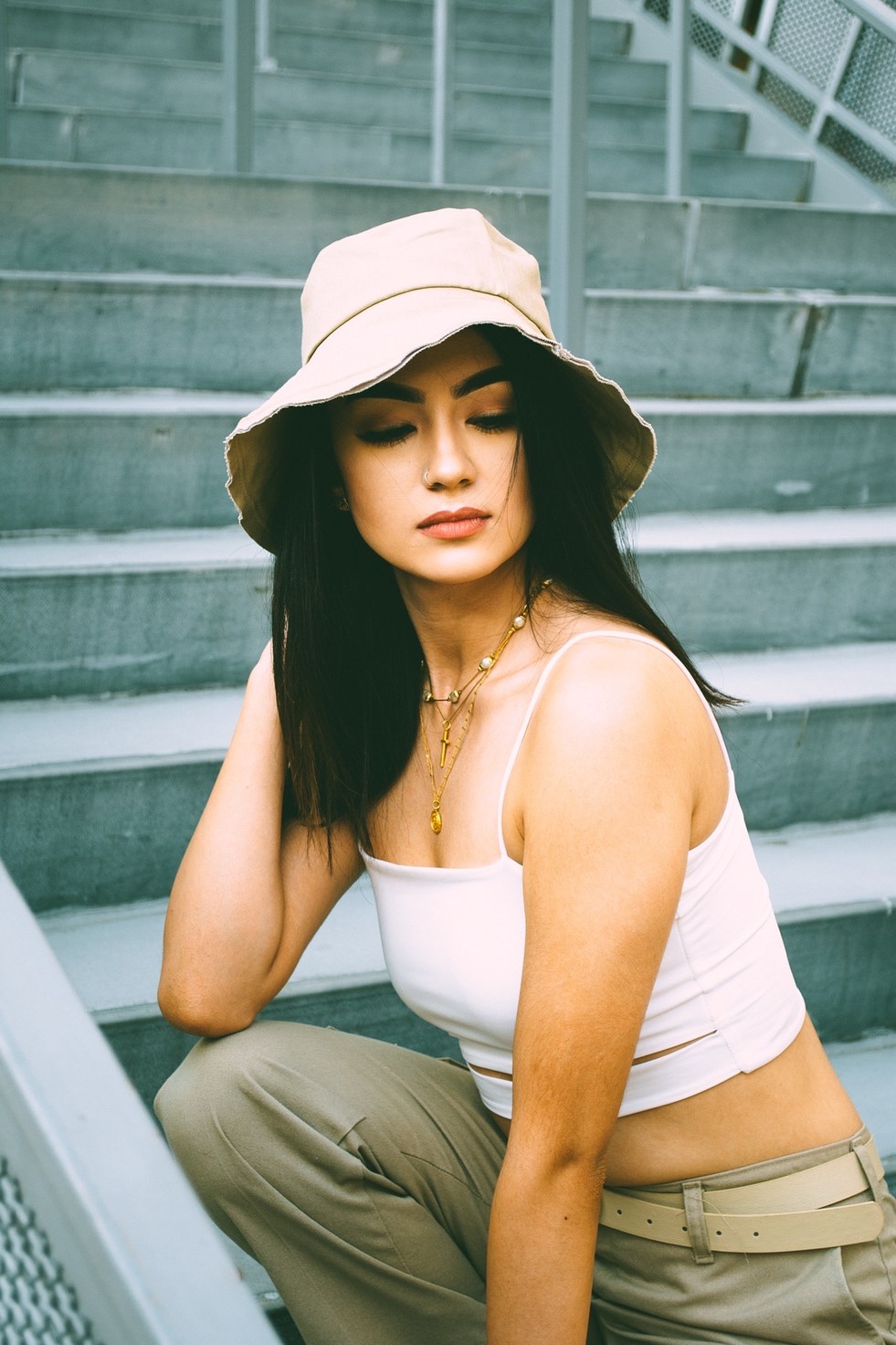 A close portrait in a bucket hat on concrete steps, eyes lowered, hand in the hair — from the BALENCI sequence