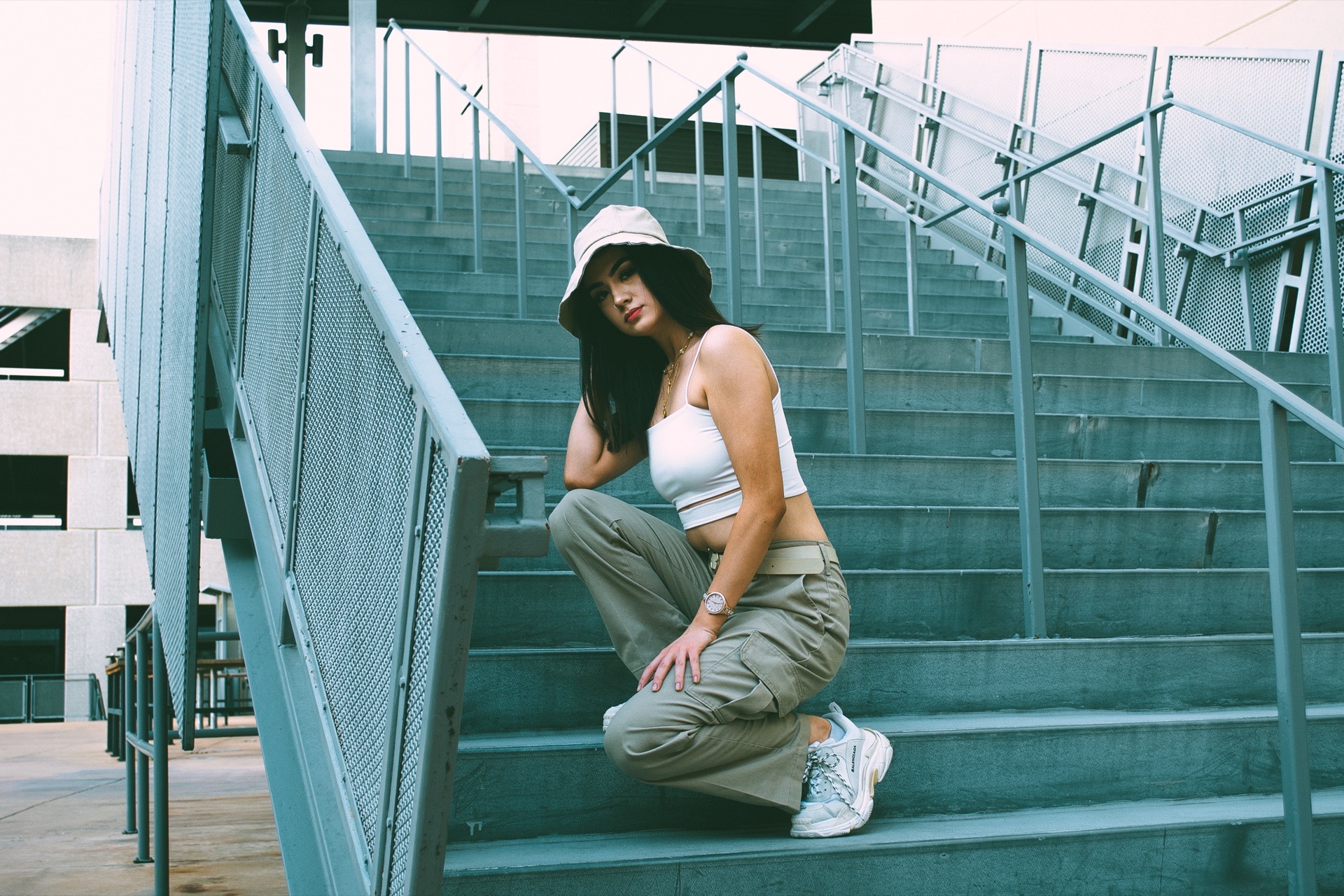 Crouched mid-stairway on wide steel-railed steps, in a bucket hat and khaki cargos — from the BALENCI sequence