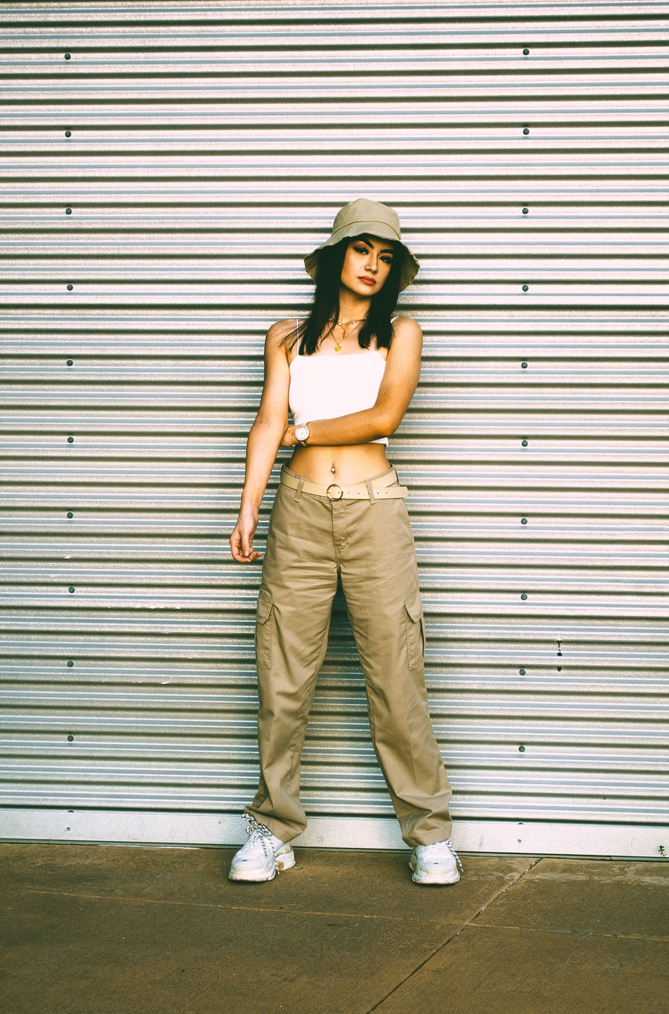 Full length against a corrugated metal wall in khaki cargos and white sneakers, one arm folded — from the BALENCI sequence