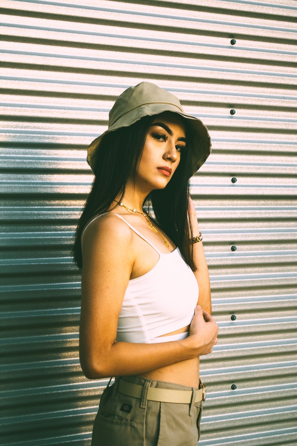A side-turned portrait in warm raking light against ribbed metal, gold chains at the neck — from the BALENCI sequence