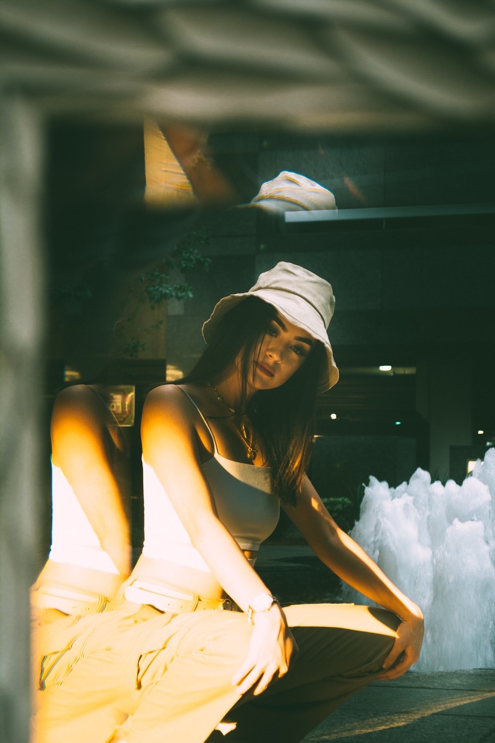 A crouched figure in amber sunlight, doubled by a glass reflection, beside a white fountain — from the BALENCI sequence
