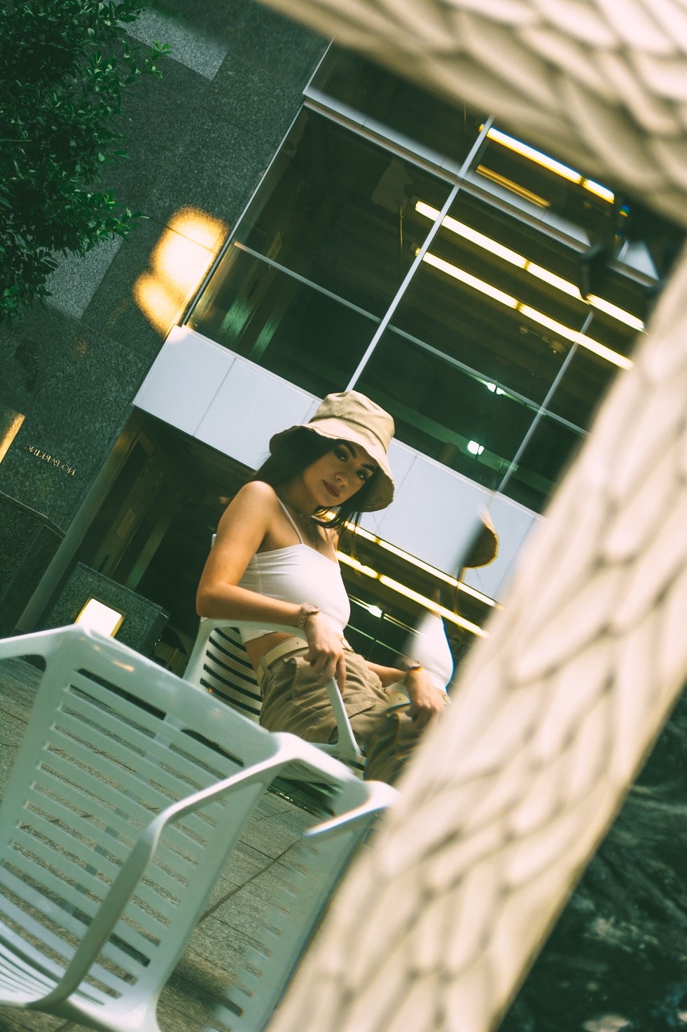 Seated on a white patio chair, seen at a tilt past a blurred carved frame — from the BALENCI sequence