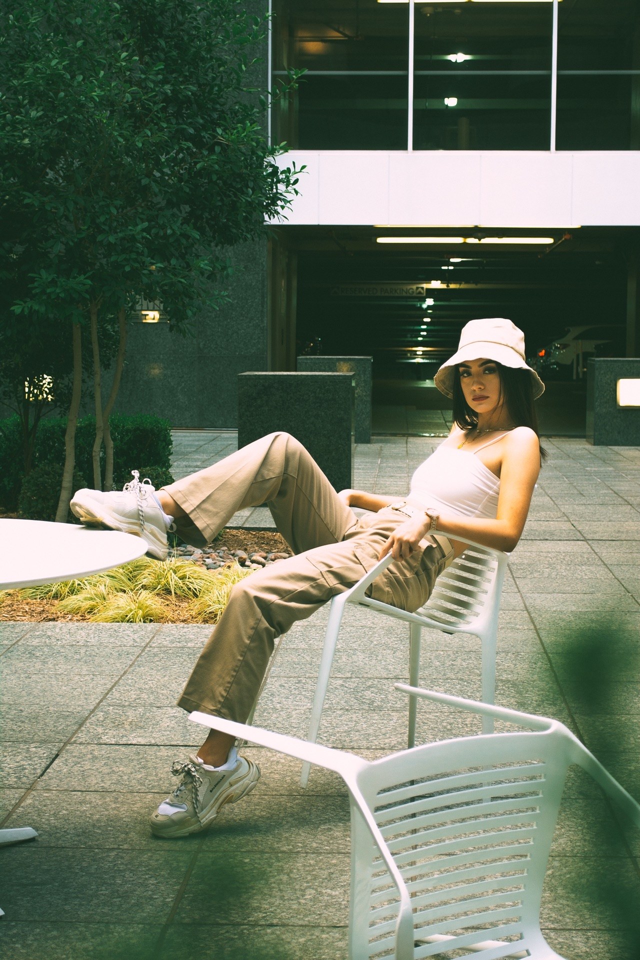 Leaning back in a white patio chair, one sneaker on a table, before a parking garage — from the BALENCI sequence