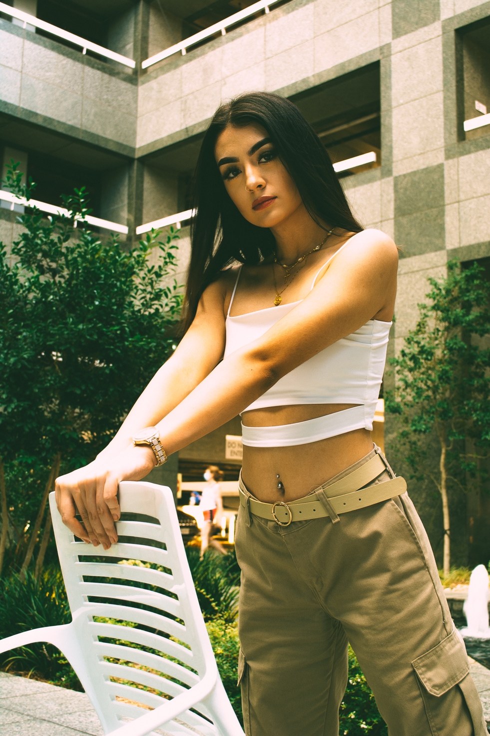 A low-angle portrait gripping the back of a white chair in a granite courtyard — from the BALENCI sequence