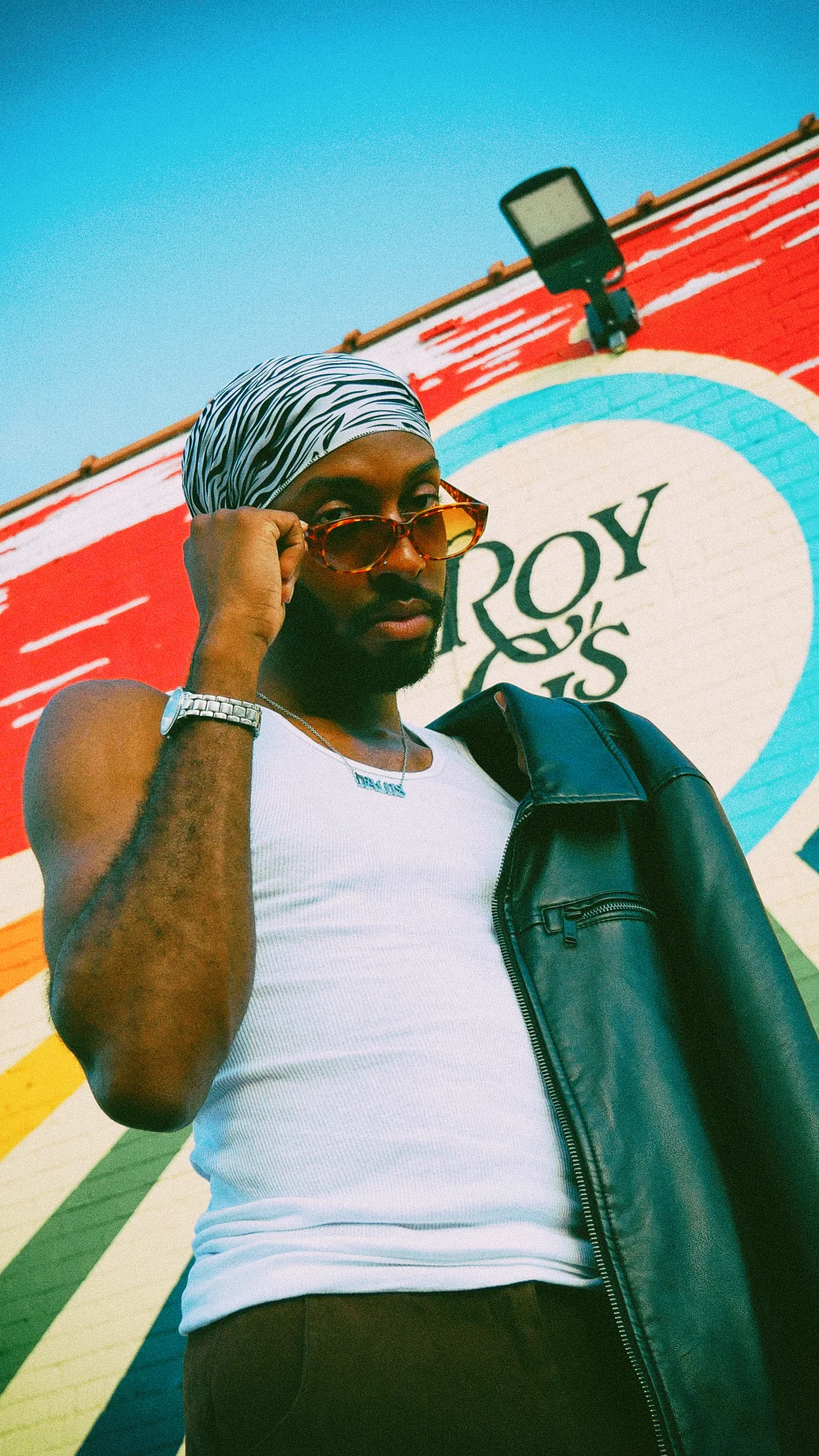 A man in a zebra-print durag lowering tortoiseshell sunglasses, a leather jacket over one shoulder, against a painted brick wall — Icon study