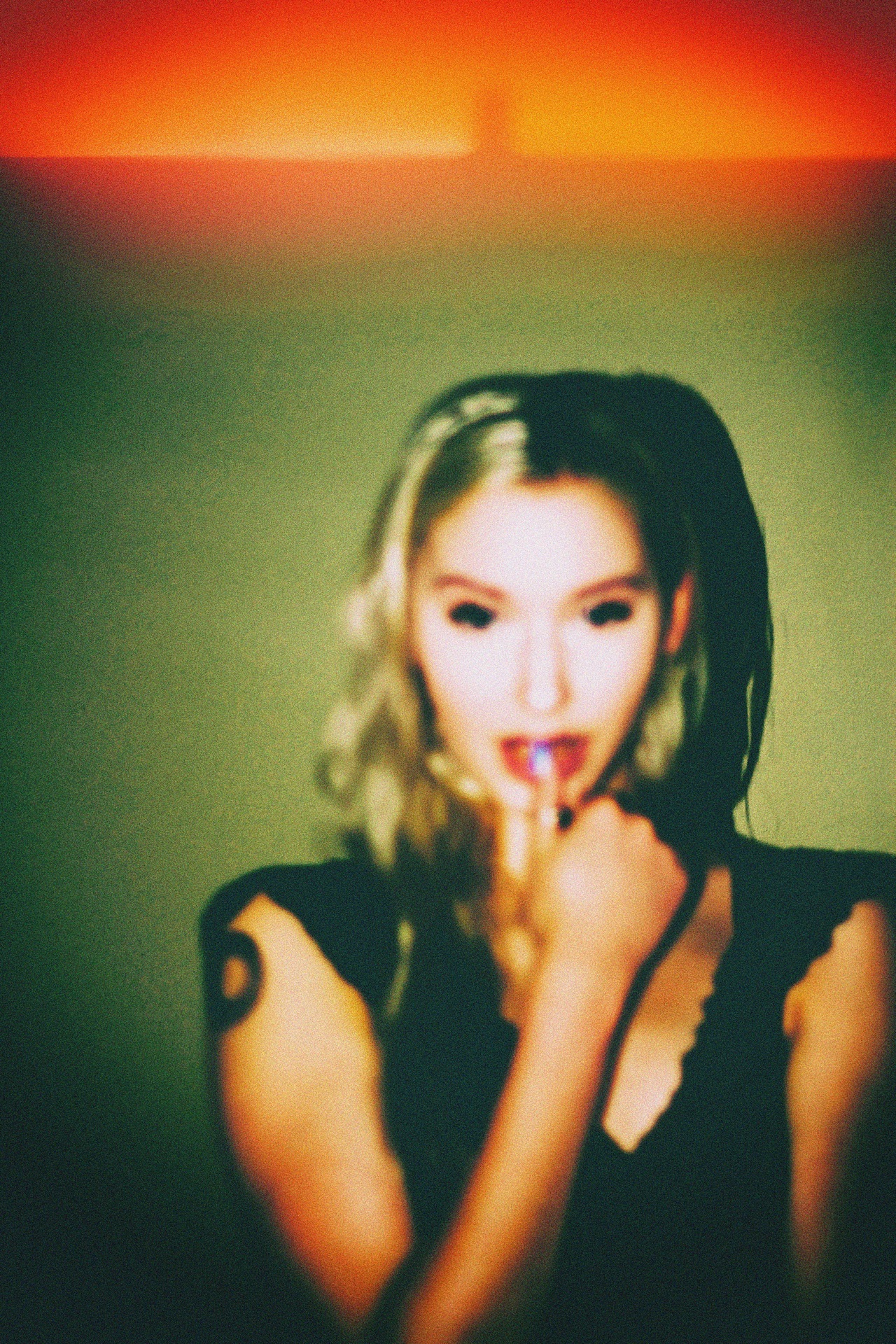 A blonde portrait in a black lace top, finger at the lips, against green and orange — from Nocturnal