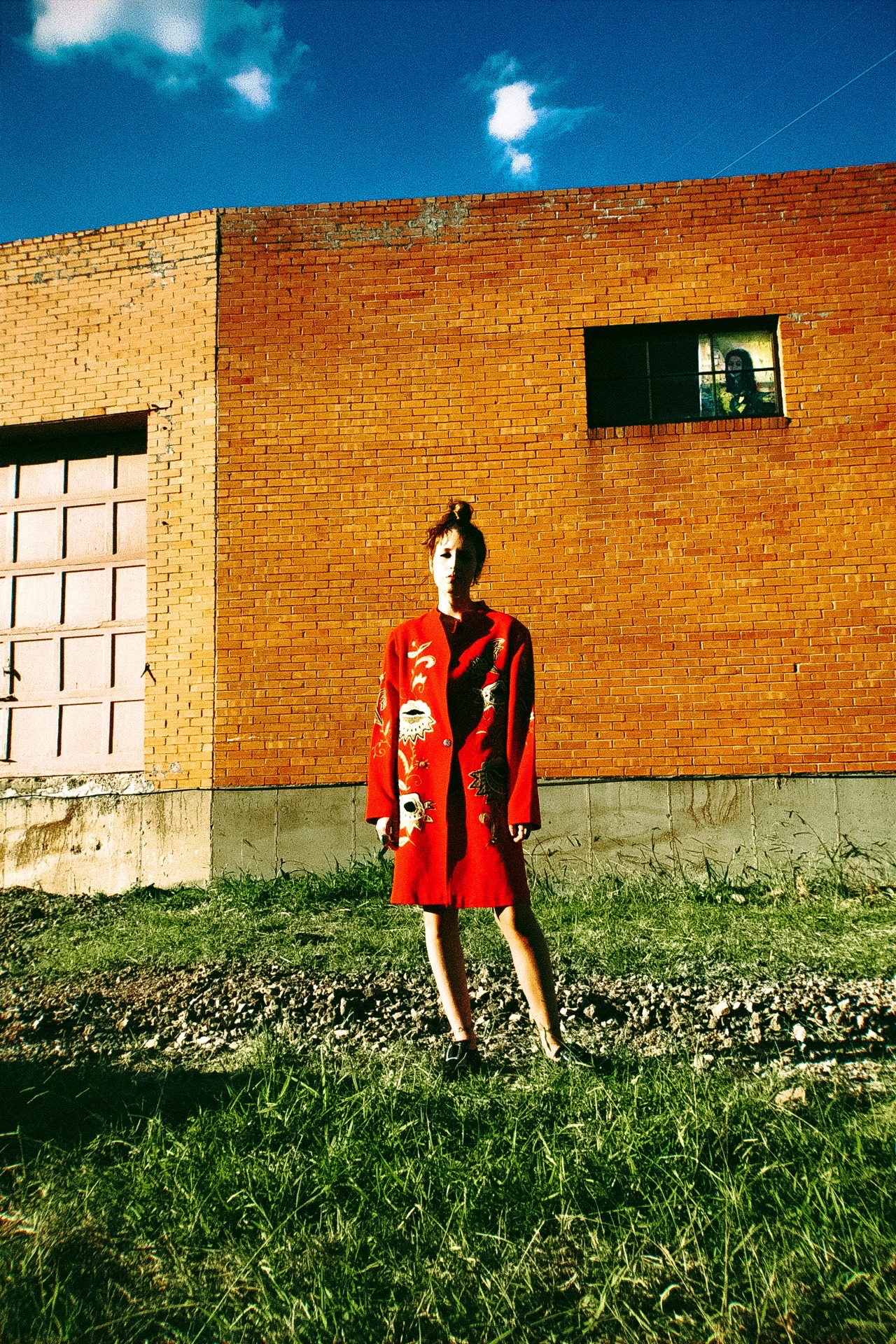 A figure in a sculptural red coat against civic brick — the Rouge study