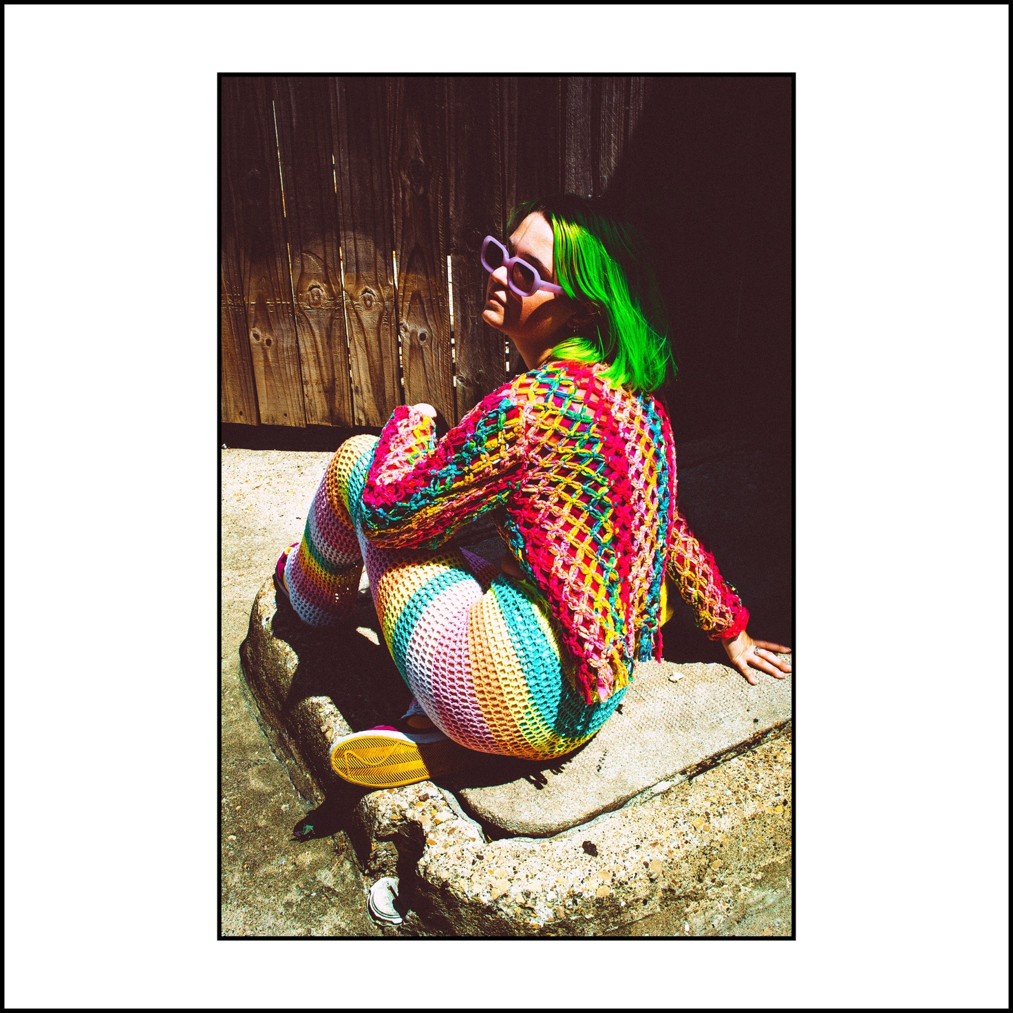 On a sunlit curb by a wooden fence, green hair and lilac sunglasses over rainbow crochet — from the STVPID study