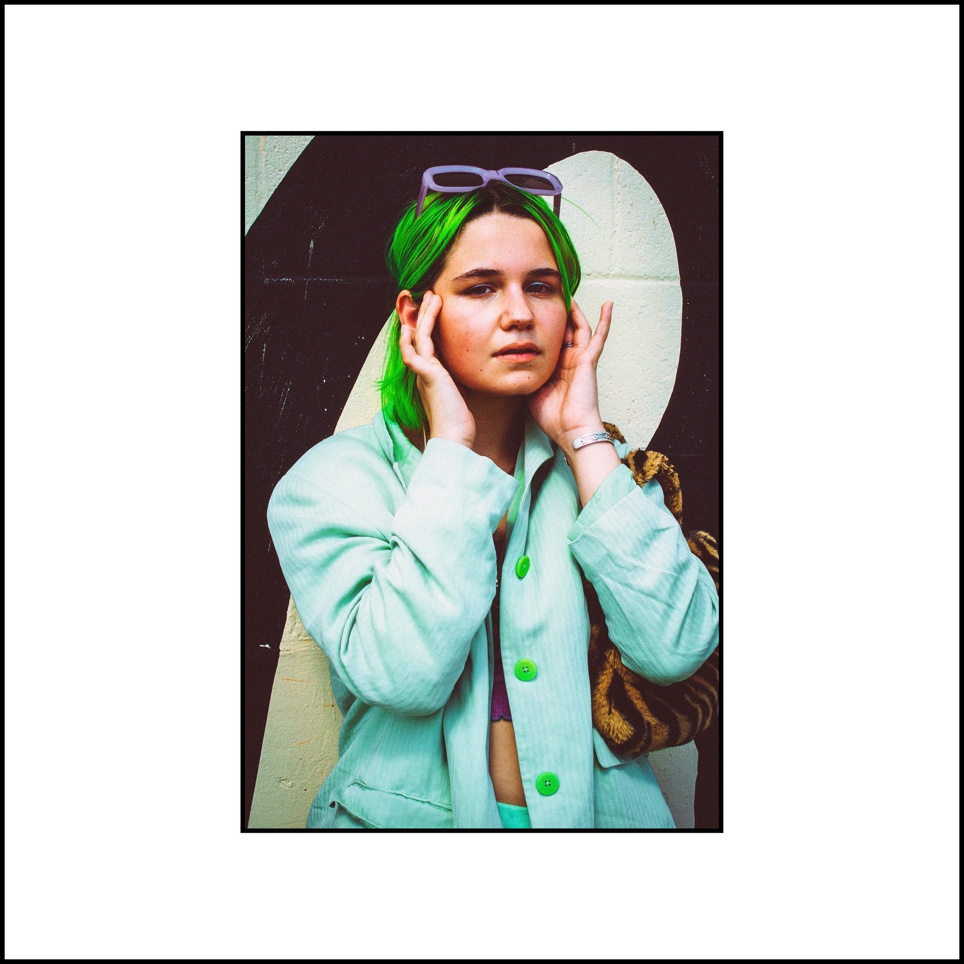Both hands framing the face, green hair and a mint jacket against a black-and-cream painted wall — from the STVPID study