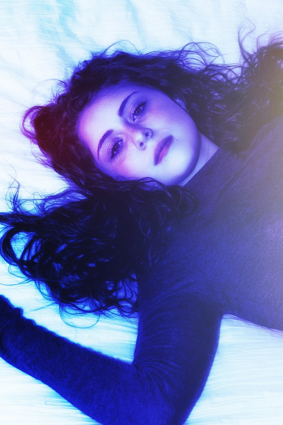A reclining blue-lit portrait with hair spread across pale bedding